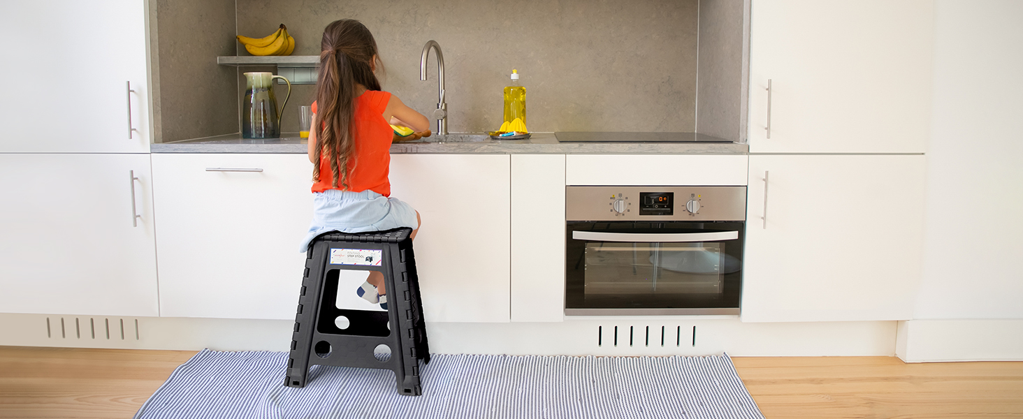Intérieur de cuisine blanc moderne avec une personne debout sur un marchepied devant l'évier, des armoires blanches, un four encastré et un tapis de sol rayé.